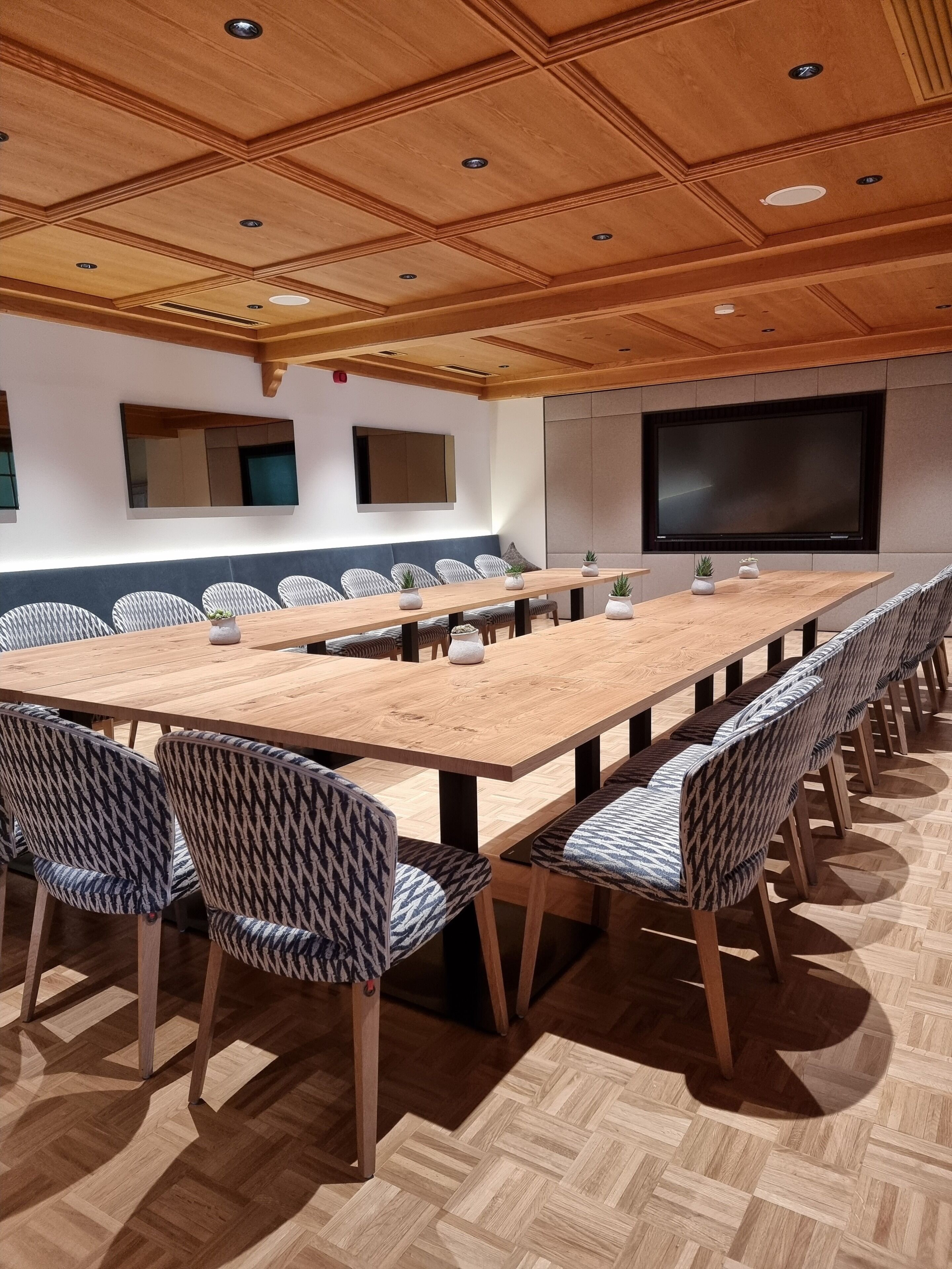 Ett konferensrum med ett långt bord, mönstrade stolar, en stor skärm och ett träpanelat tak.