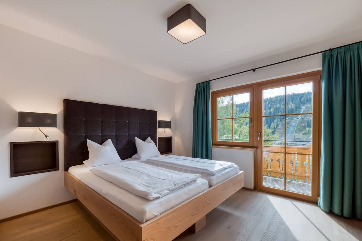 Un dormitorio moderno con una cama grande, dos mesitas de noche, un cabecero y un ventanal con vistas a un bosque y una carretera.
