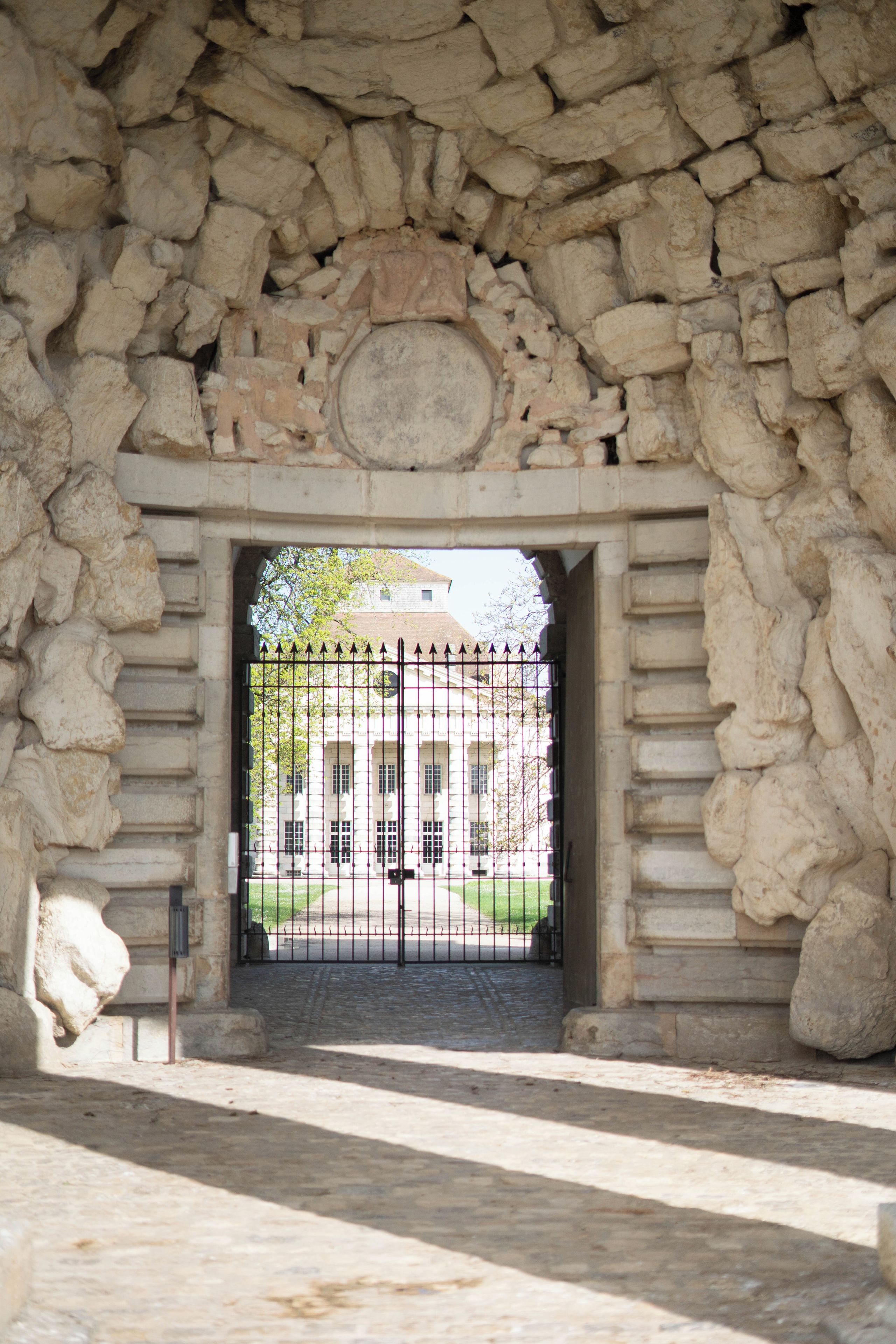Un arco de piedra que da acceso a una entrada con puerta y un edificio al fondo.