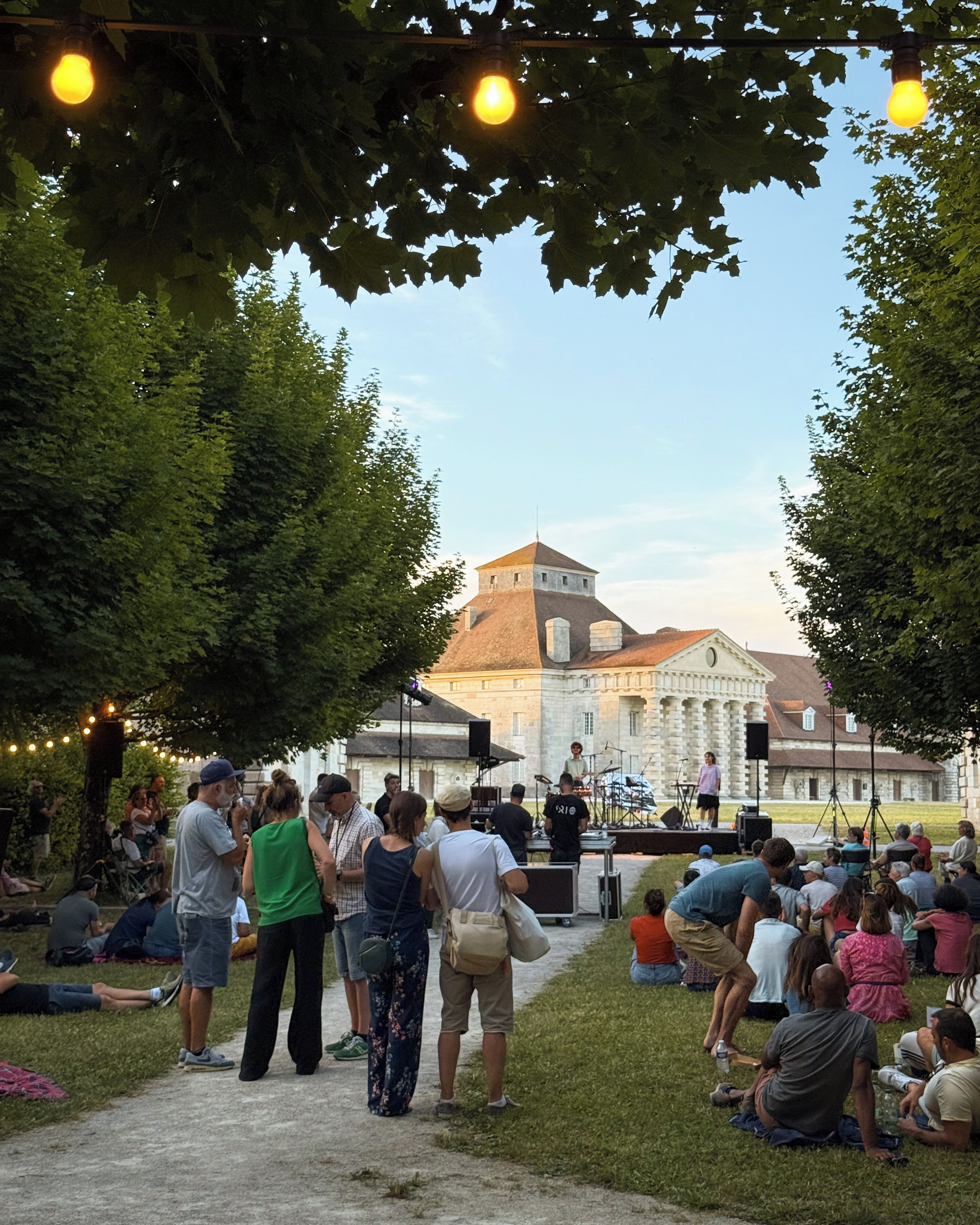 Een openluchtbijeenkomst met een podium en publiek, omringd door bomen en een gebouw met een opvallend dak.