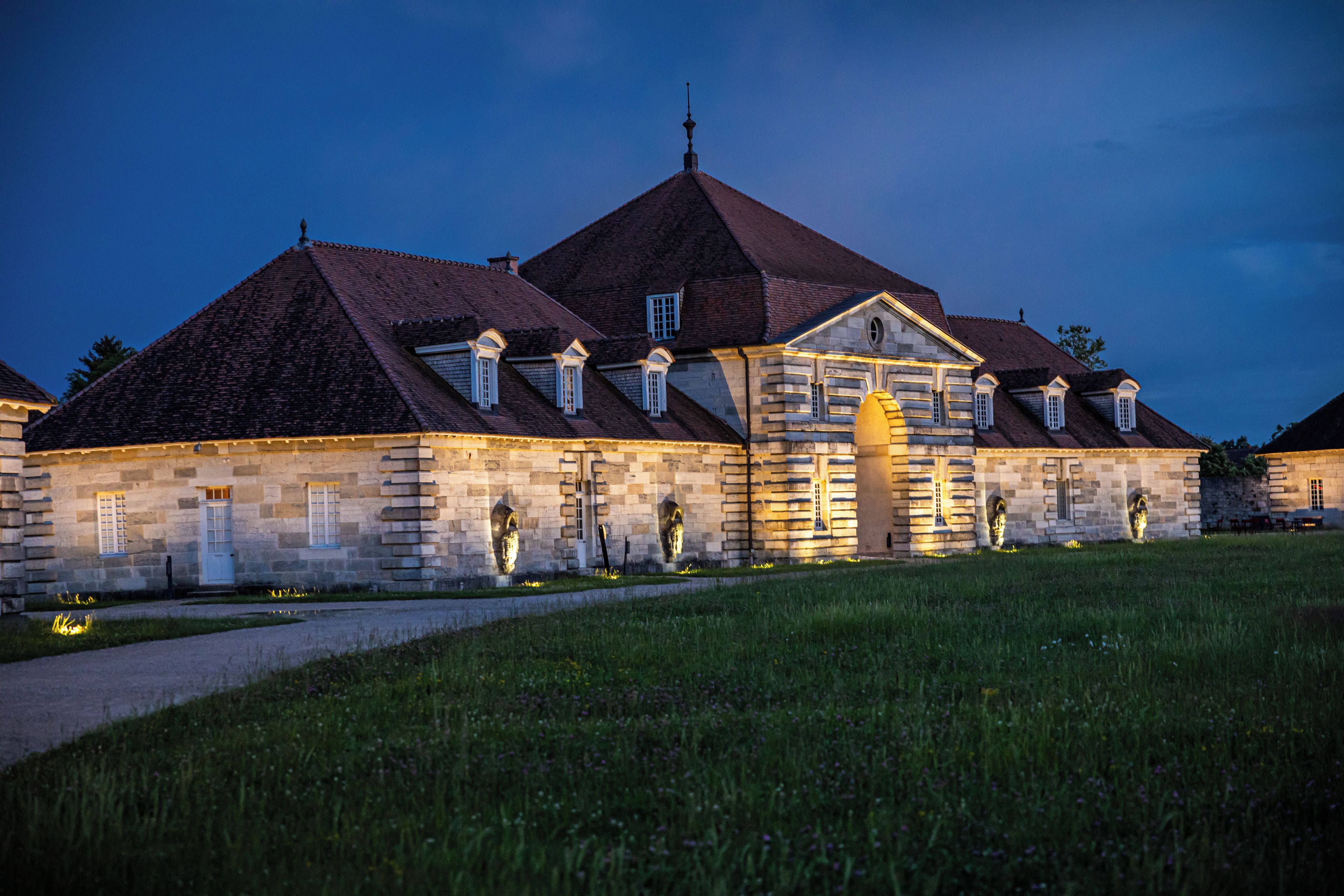 A historic stone building with multiple gabled roofs, a central entrance, and a well-maintained lawn.