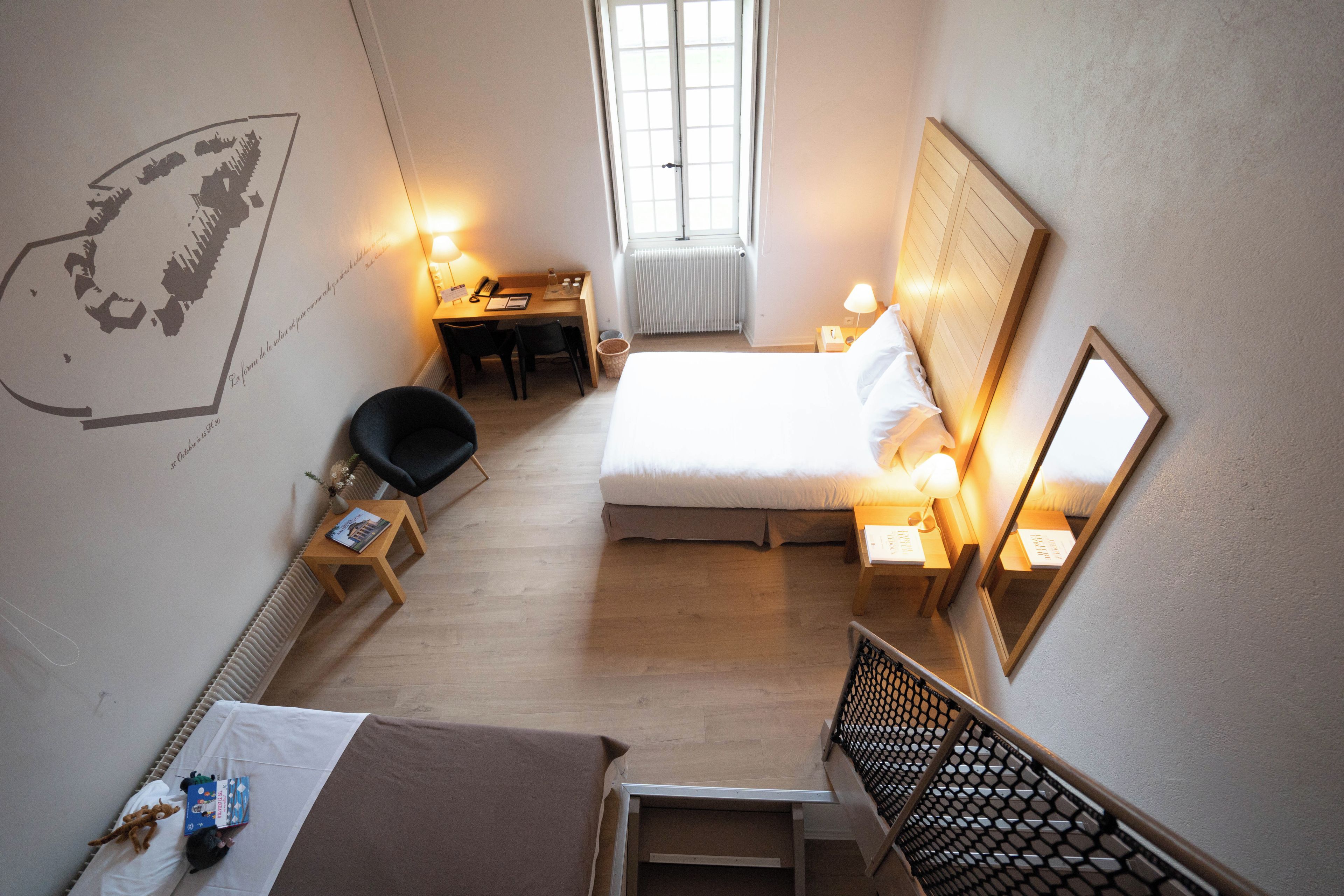 A hotel room with a bed, a desk, a chair, a small table, a window, and a wall with a map.