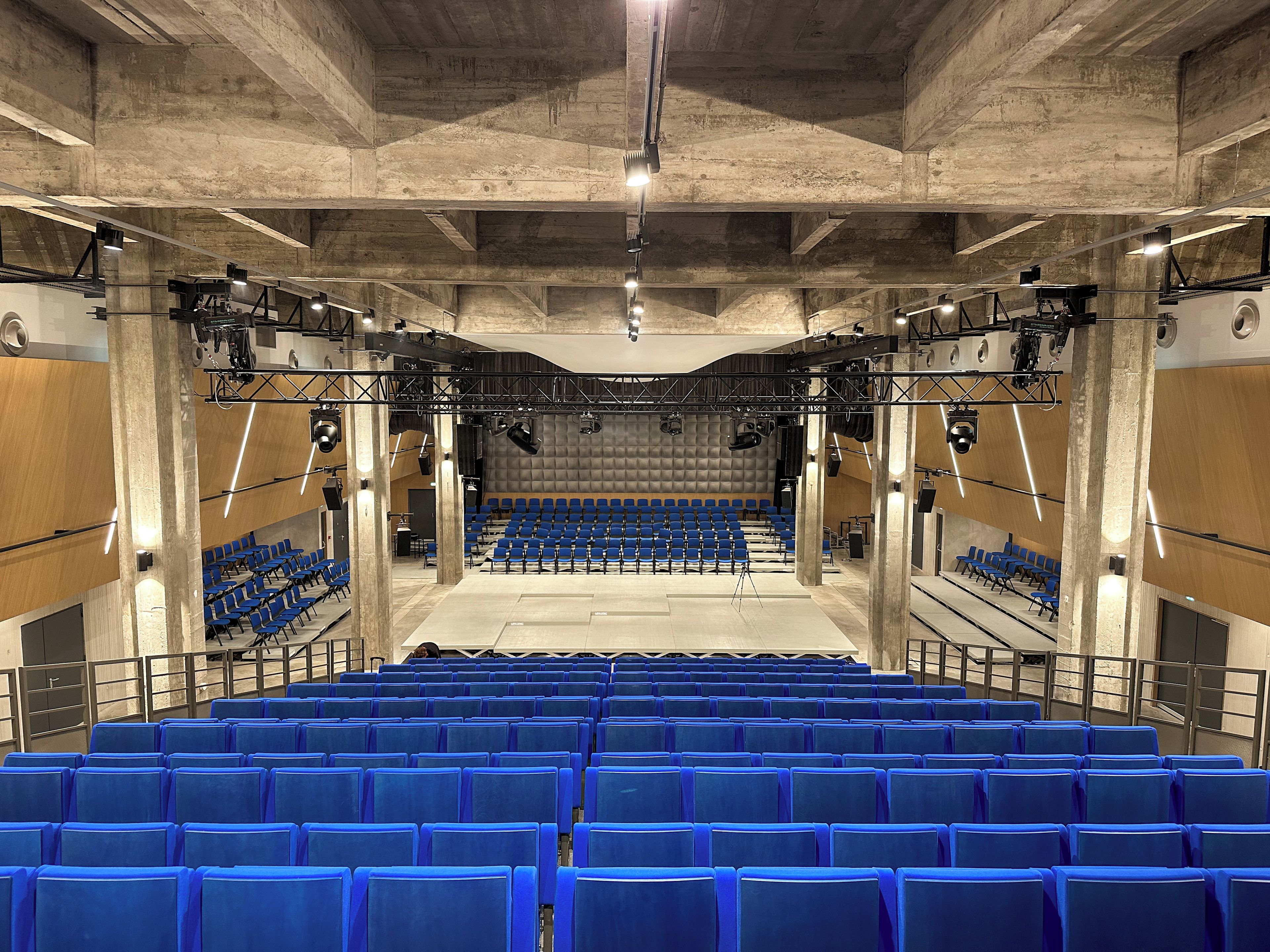 An auditorium with rows of blue chairs, a stage with lighting equipment, and a concrete ceiling.