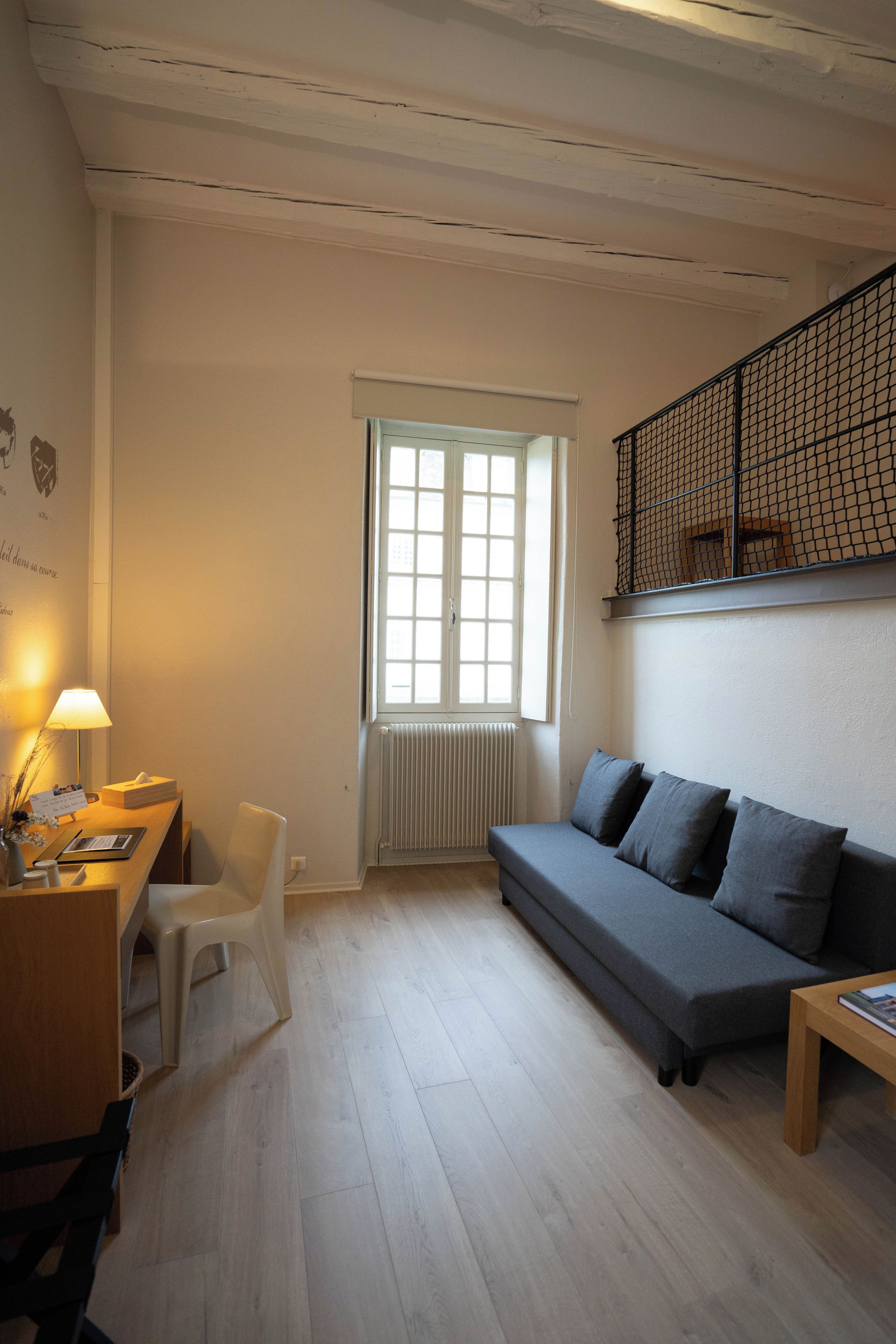 A room with a wooden floor, a grey sofa, a white chair, a desk with a lamp, and a window with blinds.