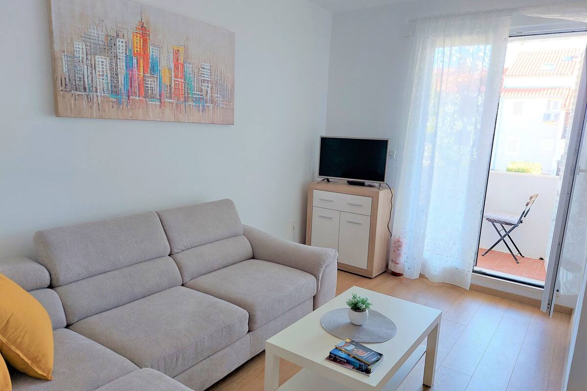 A living room with a sofa, a coffee table, a TV on a cabinet, and a balcony with a chair.