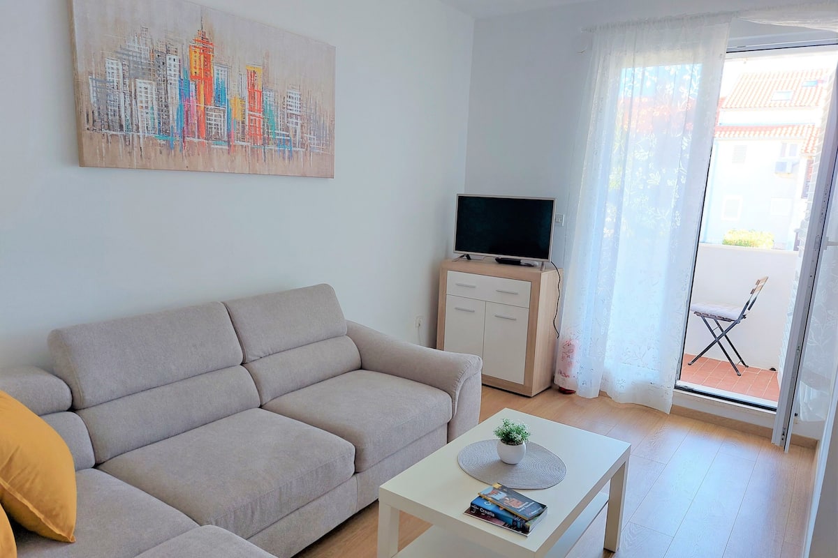 A living room with a sofa, a coffee table, a TV on a cabinet, and a balcony with a chair.
