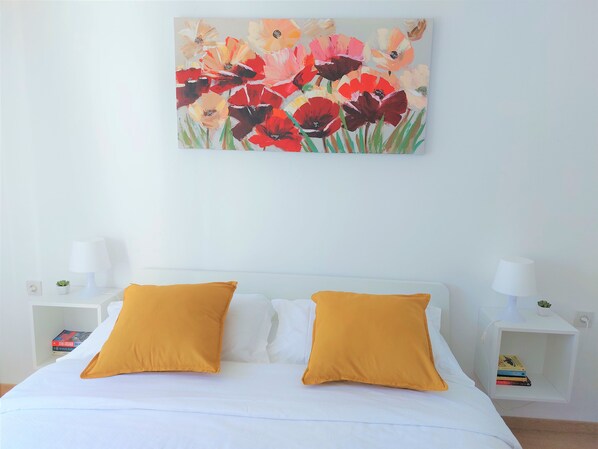 A bedroom with a bed, two yellow pillows, a white headboard, a white nightstand with a lamp, and a wall-mounted painting of flowers. - Apartment Tina (Dubrovnik)