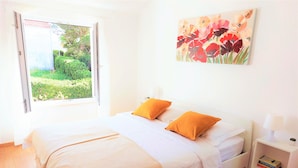 A bedroom with a bed, two pillows, a bedside table with a lamp, a small plant, and a painting on the wall.