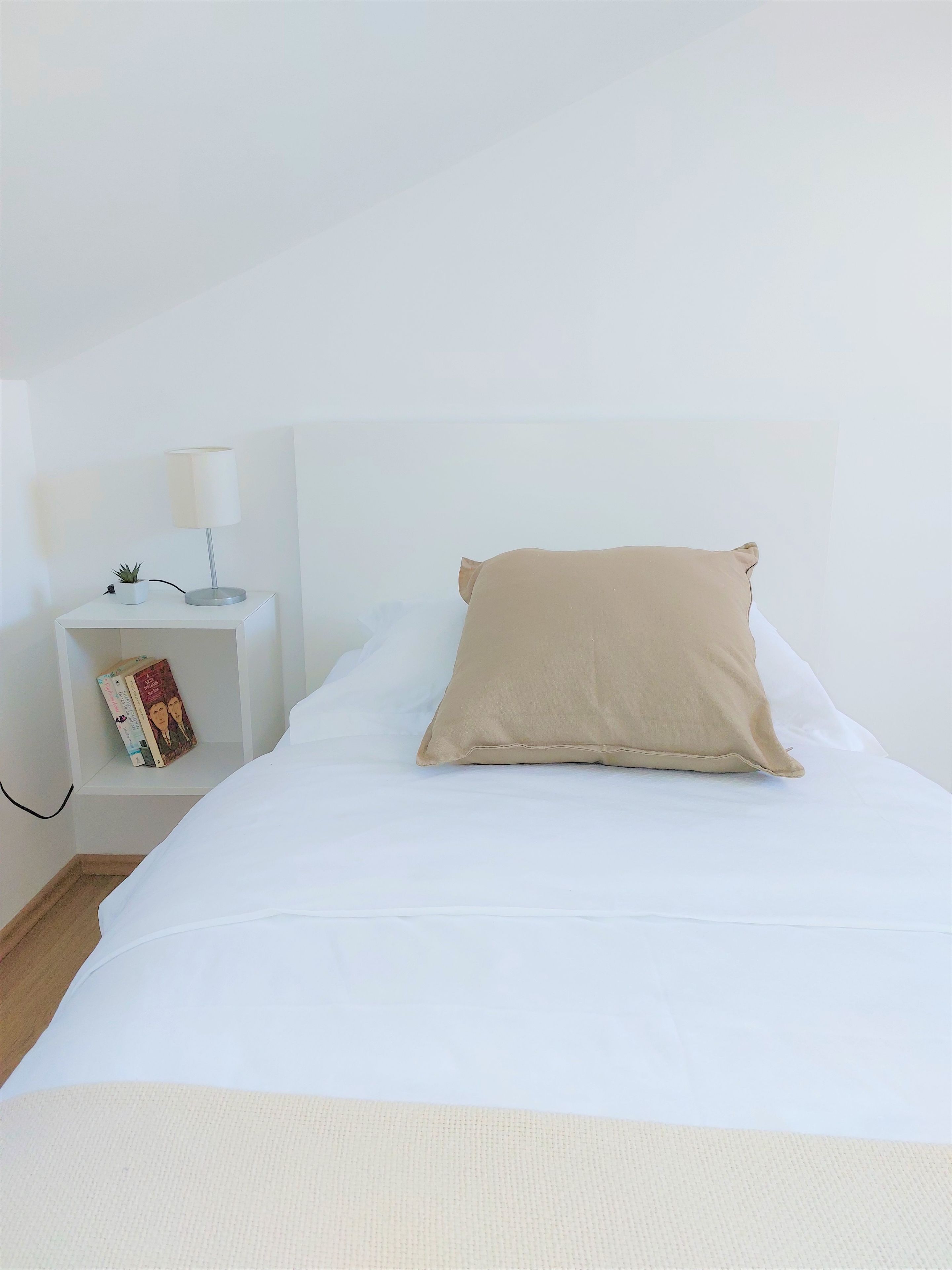 Un lit avec un matelas blanc et un oreiller beige, une table de chevet avec une lampe et un livre, et une petite plante en pot.