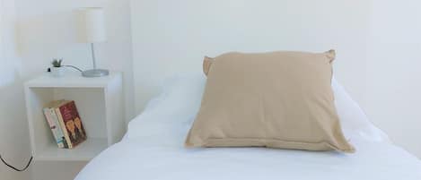 A bed with a white mattress and a beige pillow, a bedside table with a lamp and a book, and a small potted plant.