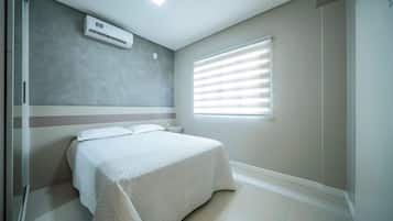Quarto de hotel com cama, janela com persianas, ar condicionado fixado na parede e luminária no teto.