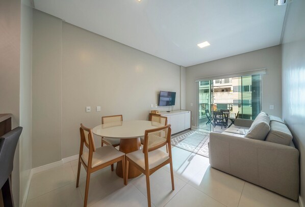 A modern living room with a sofa, a dining table with chairs, a flat-screen TV mounted on the wall, and a glass door leading to an outdoor area.