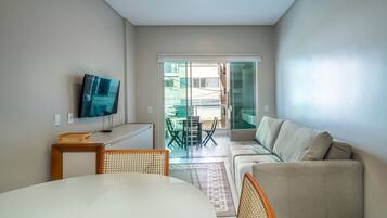 Uma sala de estar moderna com sofá, televisão na parede, mesa de jantar com cadeiras e vista para a varanda.