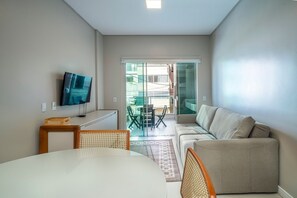 A modern living room with a sofa, a TV mounted on the wall, a dining table with chairs, and a view of the outside balcony.