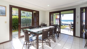 A dining area with a glass-top table and wicker chairs, large sliding glass doors leading to a balcony with a view of the ocean.