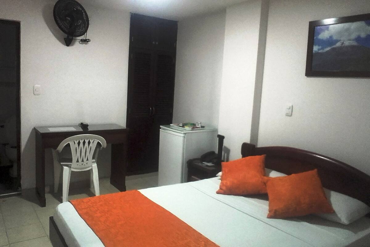 Une chambre à coucher comprenant un lit, un bureau, une chaise, une table de chevet et un ventilateur fixé au mur.
