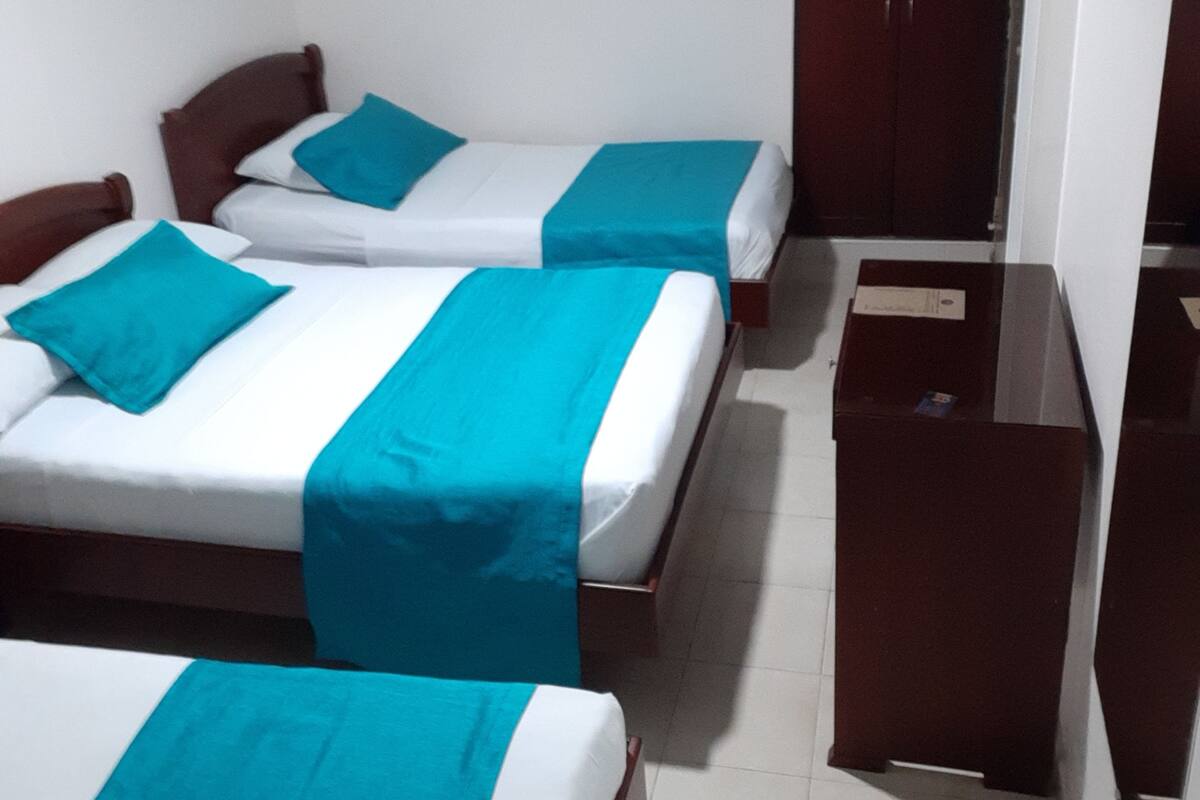 Une chambre d’hôtel avec deux lits, chacun recouvert d’une couvre-lit turquoise et de draps blancs. On y trouve une armoire en bois et une petite table sur laquelle est posée une boîte.