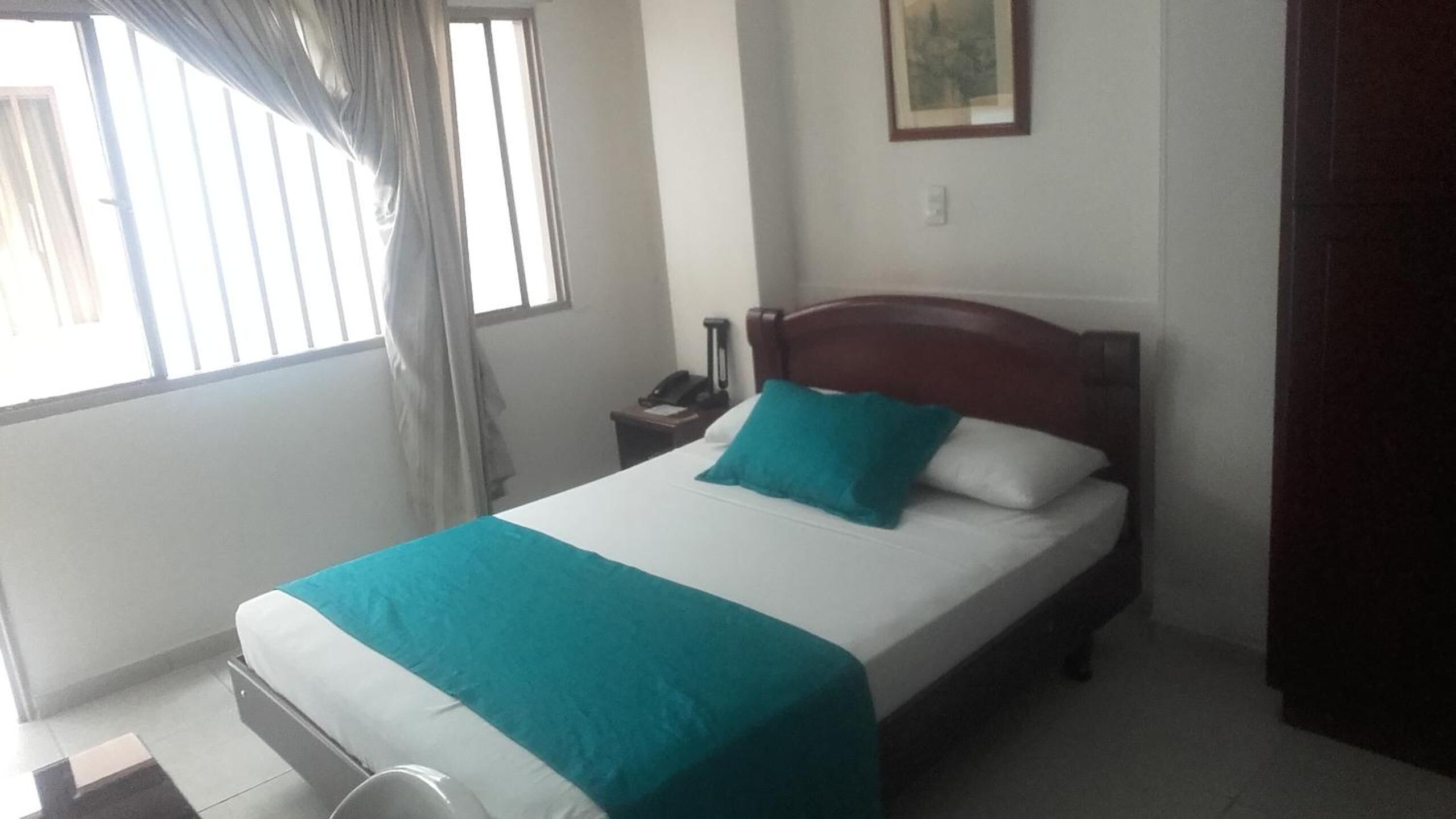 Un lit bien fait, avec une couvre-lit et des oreillers de couleur turquoise, une tête de lit en bois, une table de chevet avec un téléphone et une fenêtre avec des rideaux.