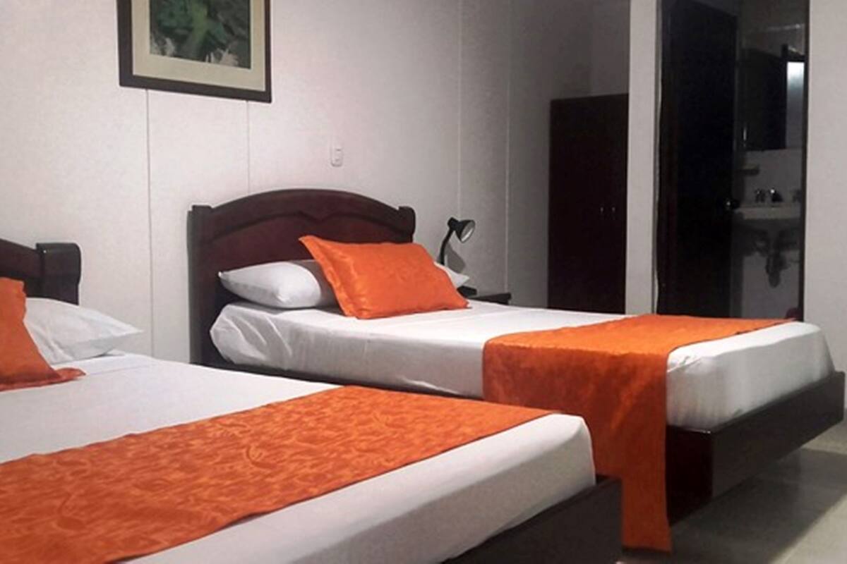 Deux lits simples avec du linge de lit blanc et des coussins décoratifs orange, une tête de lit en bois et un tableau encadré au mur.