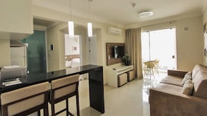 A modern living room with a sofa, a dining area with a table and chairs, a kitchen with a black countertop, and a TV mounted on the wall. - Aluguel Apartamento 1 quarto Summer Beach 076A (Bombinhas)