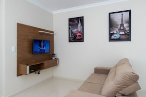 Un salon avec un téléviseur à écran plat fixé sur un panneau en bois, un canapé beige et deux tableaux encadrés représentant des monuments célèbres.