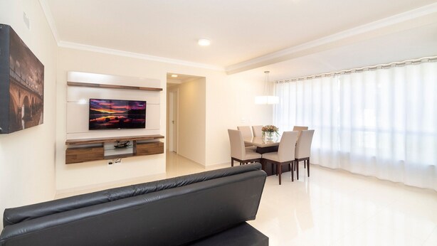 Una sala moderna con un sofá negro, un televisor de pantalla plana montado en la pared, un comedor con mesa y sillas, y un ventanal con cortinas translúcidas.
