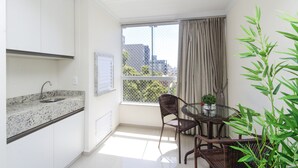 A small, bright room with a kitchen area, a dining table with chairs, and a large window with curtains.