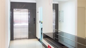 Een moderne hotelgang met een glazen lift, een telefoon en een prikbord.