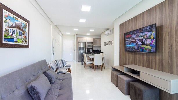 Een moderne woonkamer met een grijze bank, een televisie bevestigd aan een houten muur, en een keuken op de achtergrond.