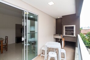 Un balcon avec une table et des tabourets blancs, une porte-fenêtre coulissante et un petit coin cuisine équipé d’un évier et d’une fenêtre.