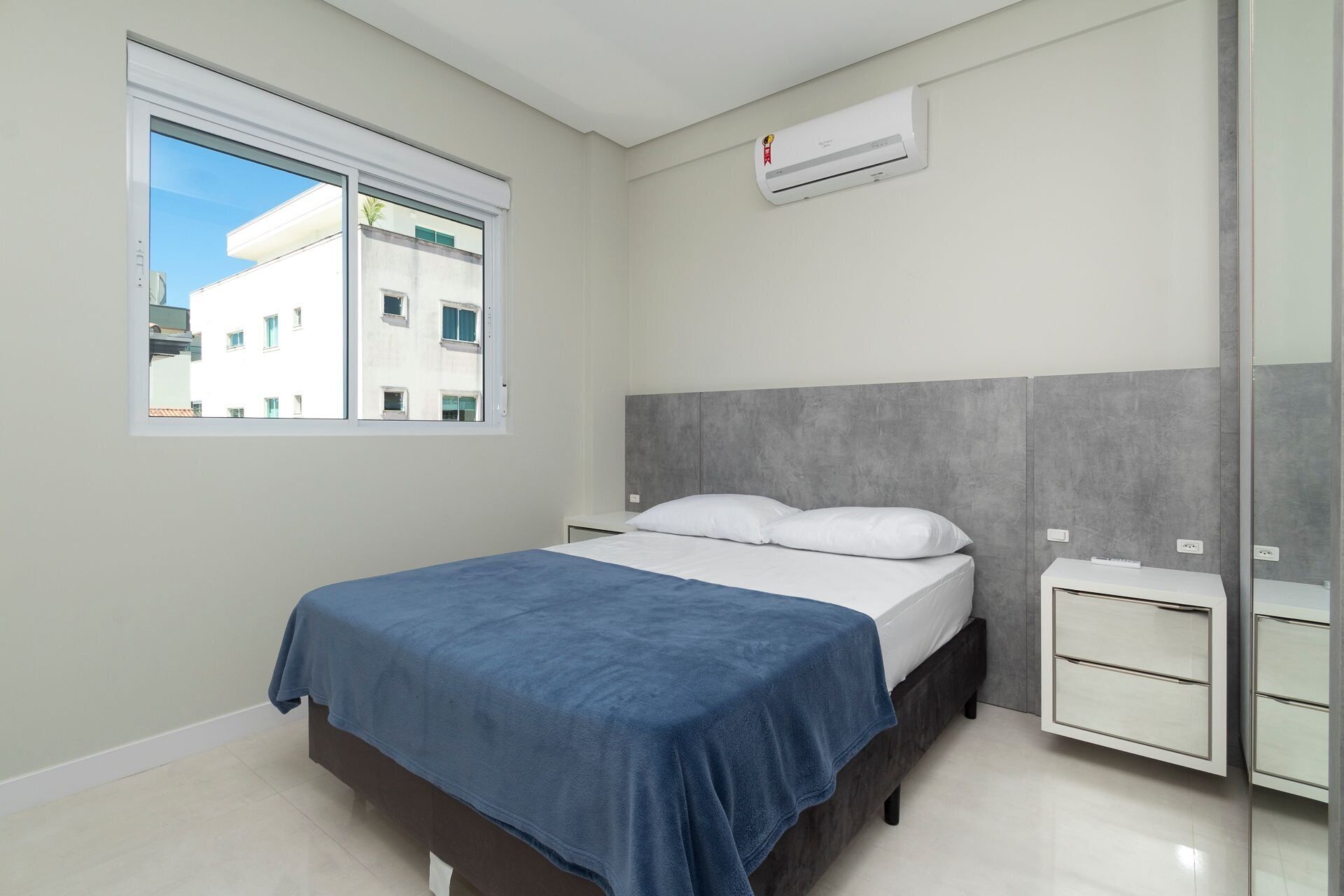 Een slaapkamer met een bed, nachtkastje, spiegel en een raam met uitzicht op gebouwen.