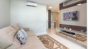 A modern living room with a beige sofa, a flat-screen TV mounted on the wall, and a built-in entertainment unit below. - Aluguel Apartamento 3 quartos 3 suites 406 (Bombinhas)