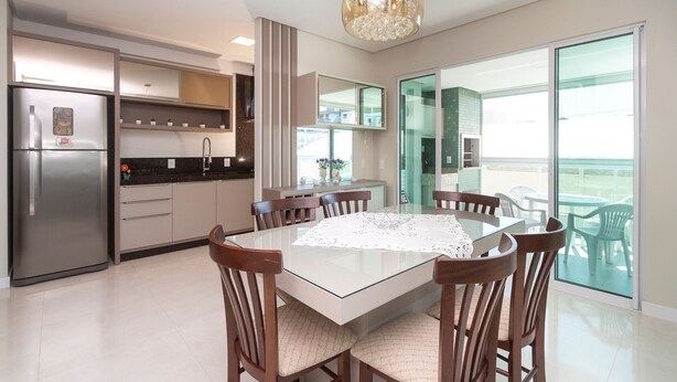 A modern kitchen with a refrigerator, stove, and sink. There is a dining table with chairs and a chandelier above. - Aluguel Apartamento 3 quartos 3 suites 406 (Bombinhas)