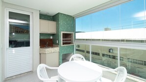 A balcony with a small kitchen area, a white table with chairs, and a large window offering a view of buildings. - Aluguel Apartamento 3 quartos 3 suites 406 (Bombinhas)