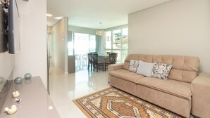 A modern living room with a beige sofa, a glass dining table, and a patterned rug. - Aluguel Apartamento 3 quartos 3 suites 406 (Bombinhas)