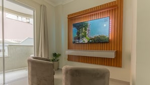 Un salon moderne avec un grand téléviseur à écran plat fixé sur un mur en panneaux de bois, deux fauteuils confortables, une plante en pot et une porte-fenêtre coulissante donnant sur un balcon extérieur.