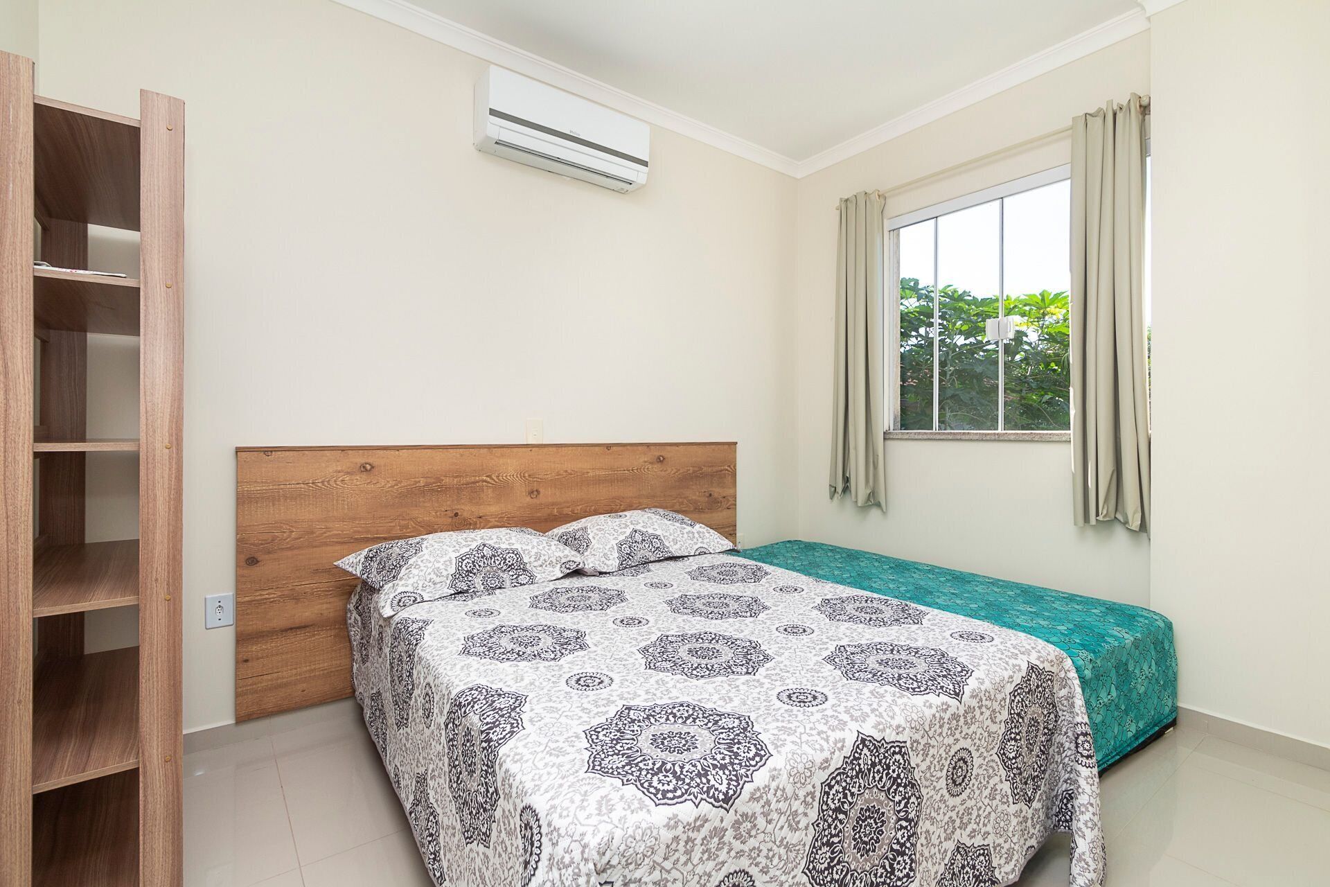 Ein Schlafzimmer mit einem Holz-Kopfteil, einem Bett mit gemusterten Betttextilien, einem Fenster mit Vorhängen und einer Klimaanlage.