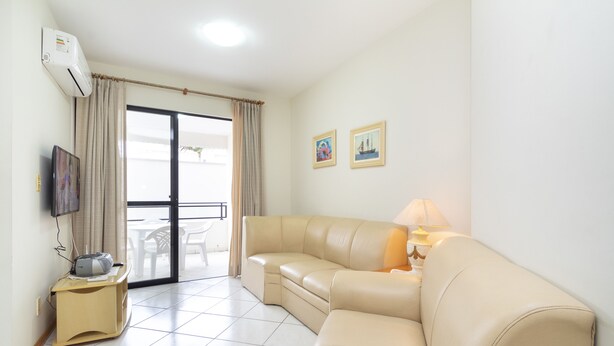 A living room with a beige sofa, a small wooden table with a lamp, a flat-screen TV mounted on the wall, and a sliding glass door leading to an outdoor area. - Aluguel Apartamento 3 quartos 1 suite 100m Mar 427 (Bombinhas)