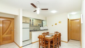 A dining area with a wooden table and chairs, a ceiling fan, and a kitchen in the background. - Aluguel Apartamento 3 quartos 1 suite 100m Mar 427 (Bombinhas)