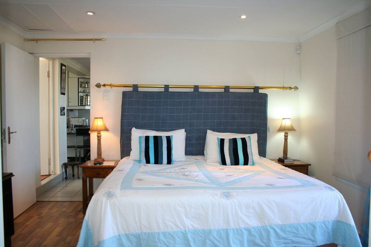 Un dormitorio con una cama grande, dos mesitas de noche con lámparas, un cabecero azul oscuro, y vistas a una cocina y un baño a través de una puerta abierta.