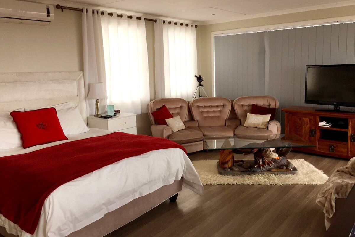 Habitación de hotel con cama, sofá, mesita de centro de vidrio, televisor y ventana con cortinas.
