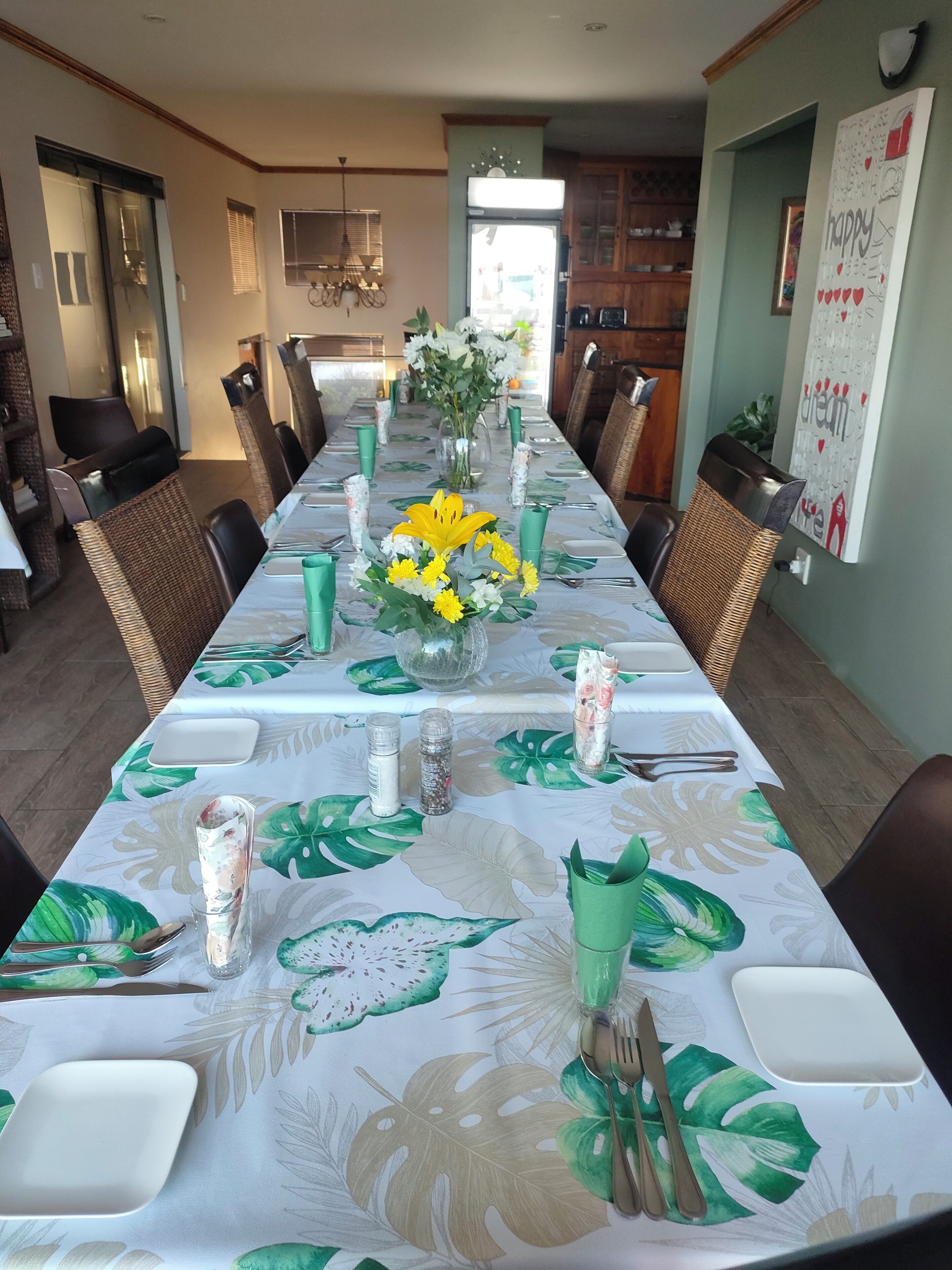 Un comedor con una larga mesa dispuesta para una comida, que incluye un mantel floral, sillas y un jarrón con flores.