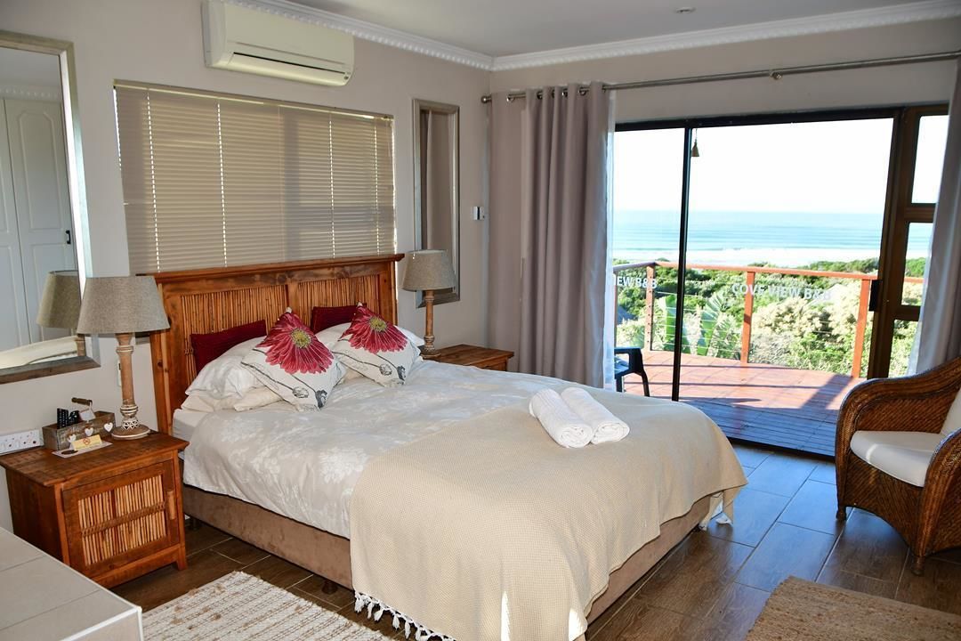 Quarto com uma cama grande, cabeceira de madeira, mesa de cabeceira com abajur e vista para o oceano através de uma porta de vidro deslizante.