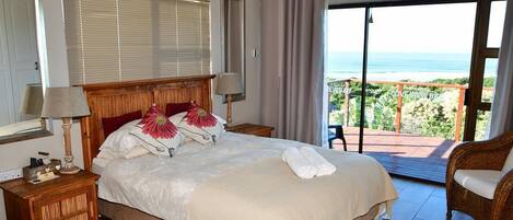 Quarto com uma cama grande, cabeceira de madeira, mesa de cabeceira com abajur e vista para o oceano através de uma porta de vidro deslizante.