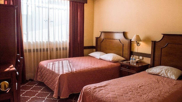 A hotel room with two beds, a nightstand, a lamp, and a window with curtains. - Hotel de Don Pedro (Guatemala City)