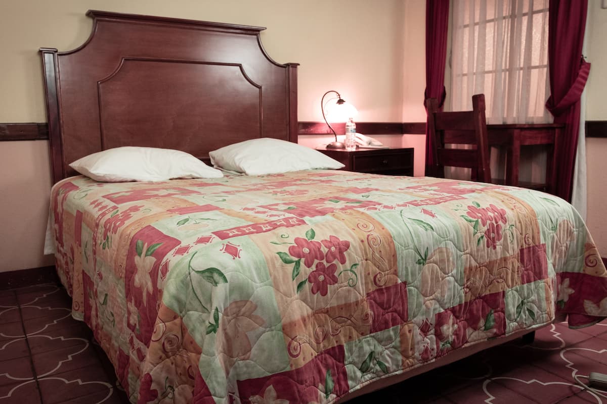 A bed with a floral quilt, a wooden headboard, a bedside lamp, and a chair next to a window with curtains.