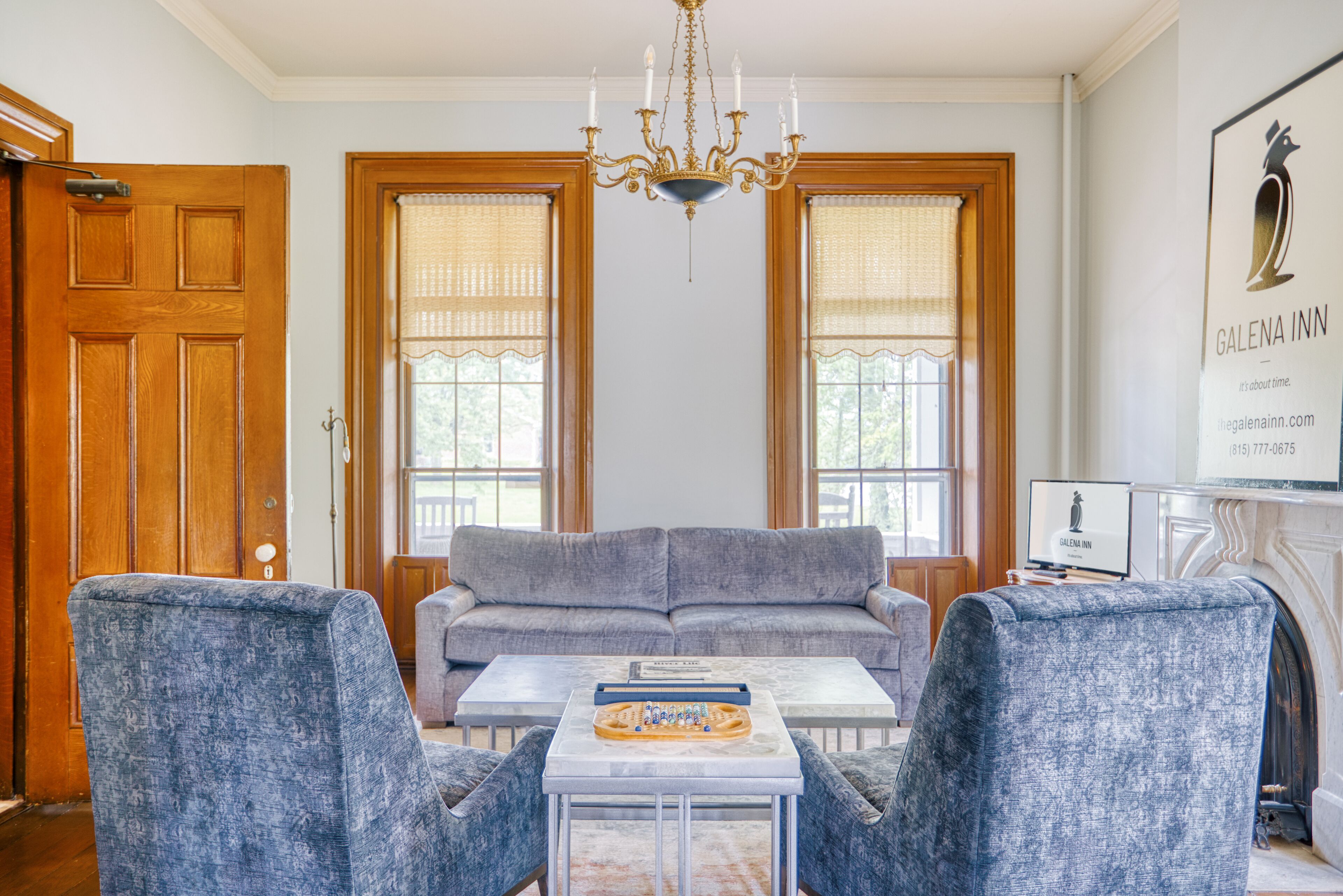Une pièce avec une porte en bois, un lustre, un canapé gris, deux fauteuils, une table basse et une cheminée.