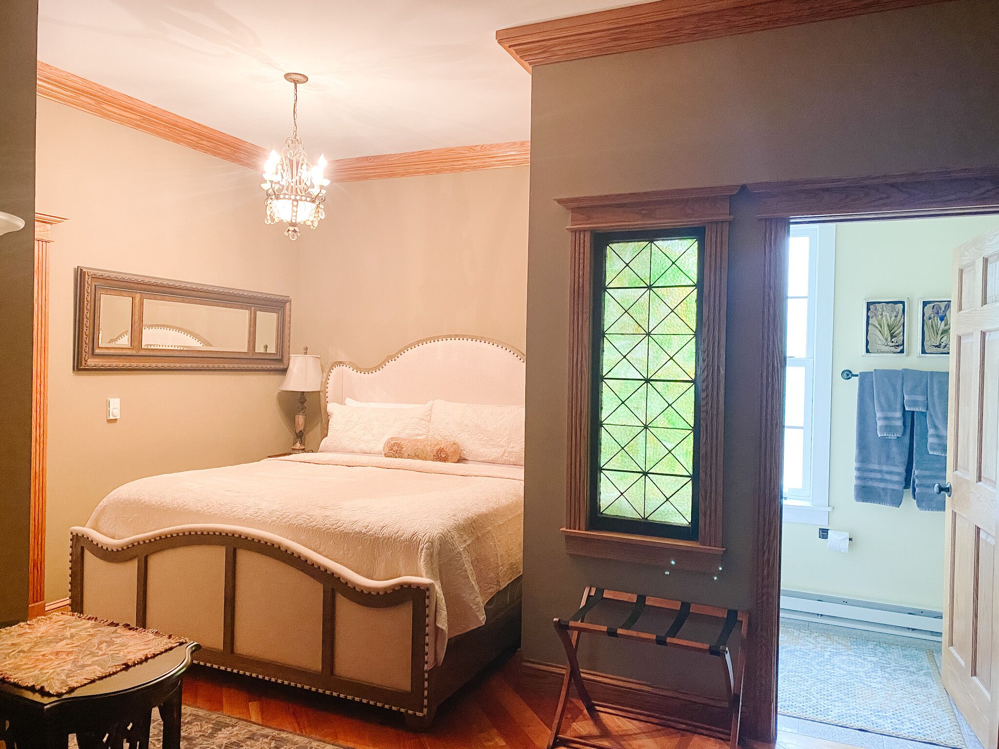 Un dormitorio con cama, una araña, un ventanal con diseño geométrico y una puerta que da al baño.