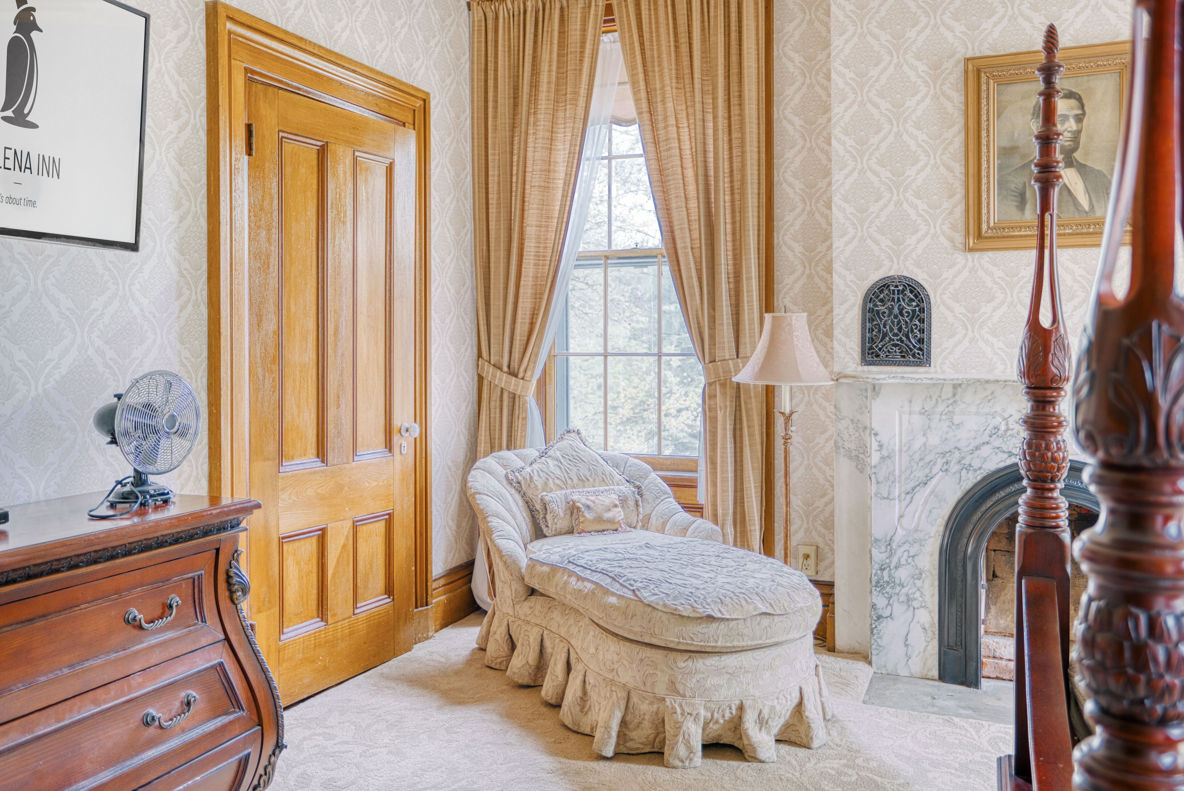 A bedroom with a wooden door, a bed with a white bedspread, a nightstand, a lamp, and a fireplace.