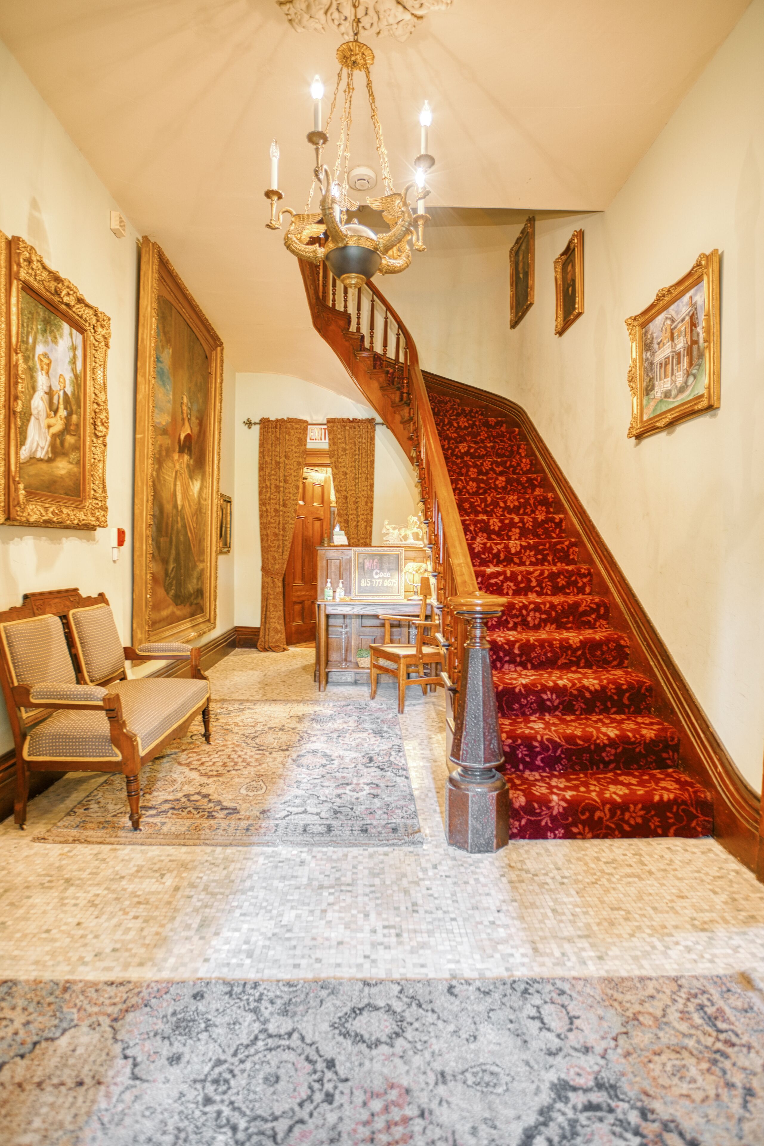 A grand staircase with ornate railings and a chandelier, leading to a room with a large painting and a wooden chair.